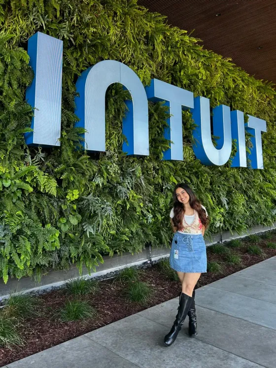 Navyaa smiling in front of the Intuit sign on a living plant wall.