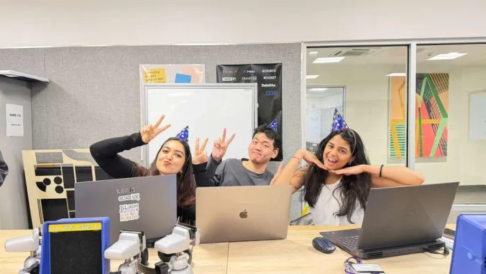 Navyaa and two teammates in party hats, making peace signs behind their laptops.